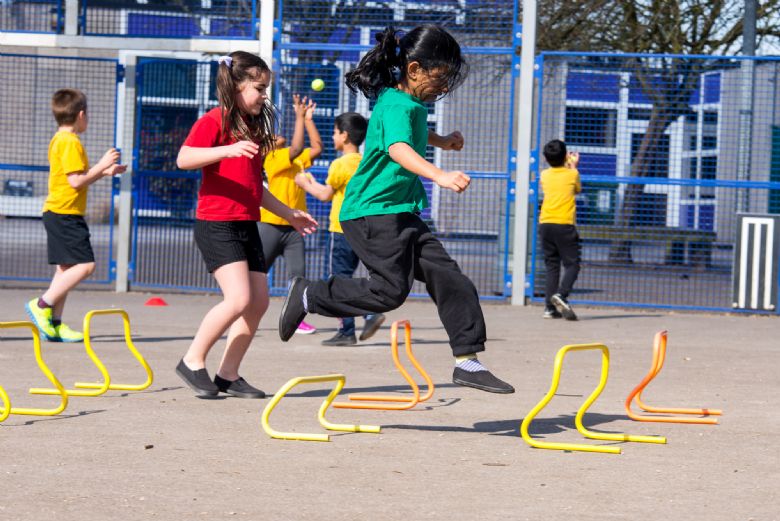 pupils jump over hurdles in a sunny playground wearing their PE uniforms